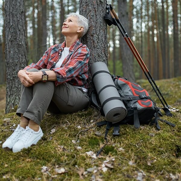 Femme âgée assise par terre, avec un sac à dos et des bâtons de randonnée à ses côtés