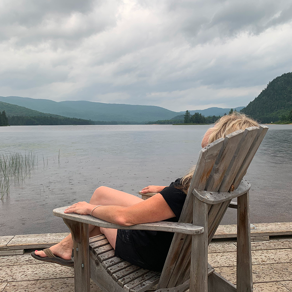 A woman sitting in a chair on a dock overlooking a serene lake, surrounded by trees and clear blue skies.