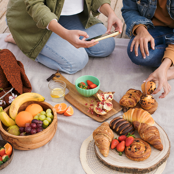 Two women enjoy a picnic on a blanket, surrounded by a table filled with various foods, celebrating family activities.
